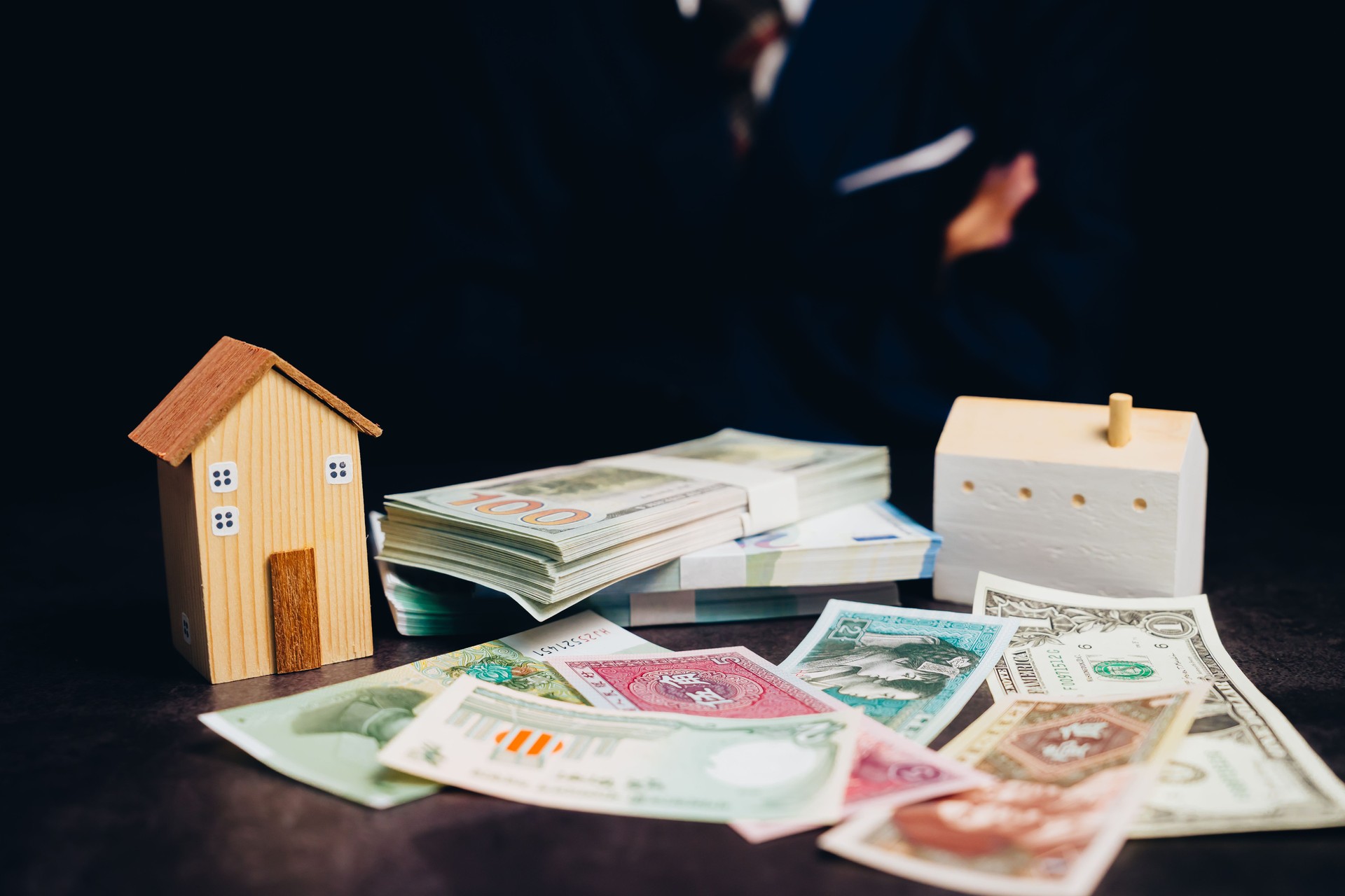 Wooden model houses beside stacks of cash and various currency notes on a dark background representing finance and investment